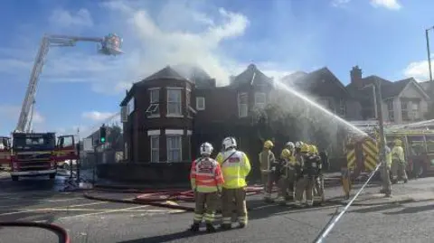 HIWFS Fire crews stood outside the home which smoke was pouring out of, with a fire engine parked to the left of the picture with a firefighter hoisted above the fire on a platform and another to the right of the picture 