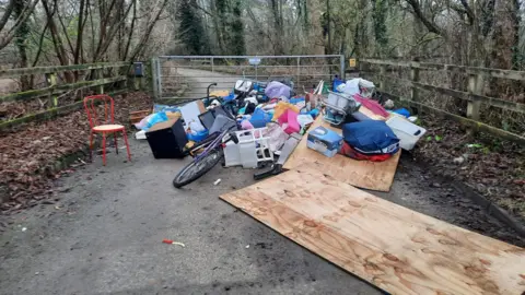 Fly-tipped rubbish, including large wooden planks, a chair, plastic bags and a bicycle, are pictured dumped on a country lane, up against a metal gate. A forest can be seen either side of the country lane. 