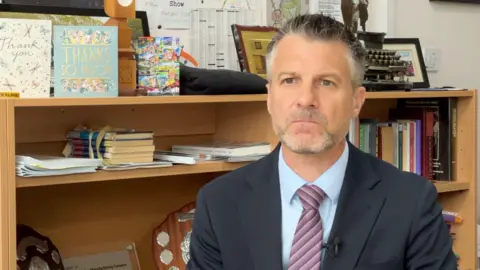 A man with grey hair and stubble, and wearing a suit and tie, sitting in an office with bookshelves behind him.