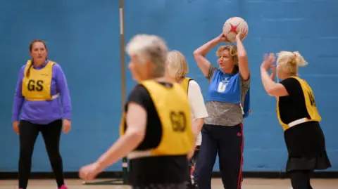 Five women in a sports hall playing netball. Three are wearing yellow bibs, one if wearing a blue bib. The one in a blue bib has her arms above her head about to throw the white ball. The wall of the sports hall is blue