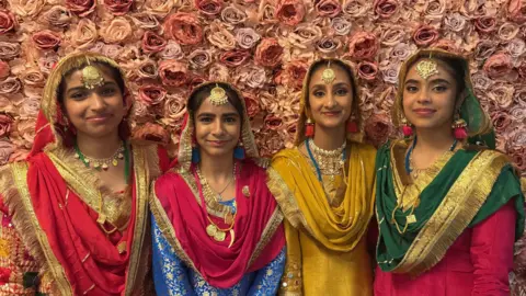 Four women with dark hair standing in front of a floral background in traditional Indian dresses made of gold, red and green fabric