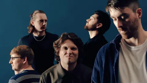 A promotional image of the band which is made up of five young men with dark hair. The man at the front on the right is looking down and is wearing a white t-shirt and blue shirt over the top. The man in the middle has longer brown hair and is scrunching up his face. On the left is a man with his back to the camera while he looks off to the to the left. He is wearing a blue striped t-shirt. Behind his is a man with brown long hair which is half tied up. He is wearing a black t-shirt and a chain necklace. To the right is a man with dark hair and a beard looking at the ceiling.