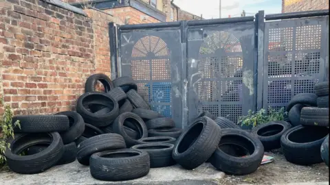 Dozens of black vehicle tyres piled beside metal alley gates between brick terraced houses.
