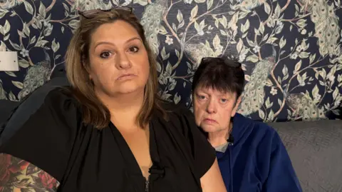 Jade Just and her mother Sarah. Jade is wearing a black top and has a large tattoo on her right arm. She has blonde-brunette hair and is wearing eye make-up with glasses on top of her head. Sarah is older and is wearing a blue hoodie. She has short, black hair and also has glasses on top of her head. 