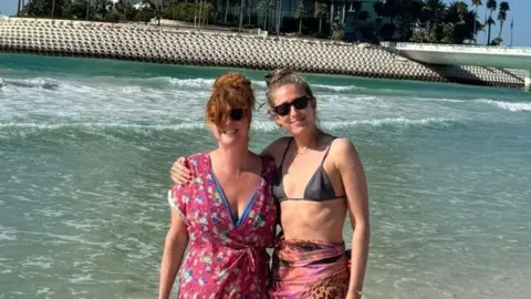 Hannah Carter Two women stand on a beach in front of a tall building shaped like a sail. The building is the Burj Al Arab. The women are wearing sunglasses. There is water in the background. 