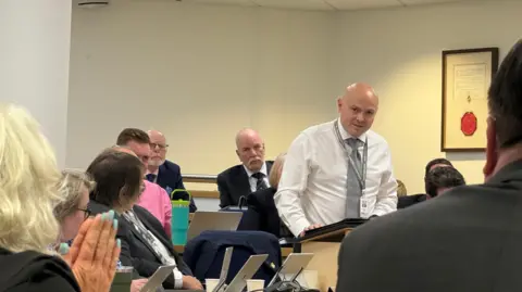 Andy Barnes talking in the council chamber, with people seated around him with laptops.