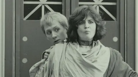 Actor Ruth Jones is pictured standing in front of another student in a performance of Casina by Plautus in 1988. She has dark hair and is wearing robes with a male actor hiding behind