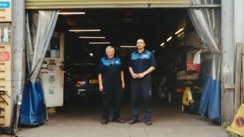 Andrew Goss Two men in work overalls stand in the entrance of a car garage.
