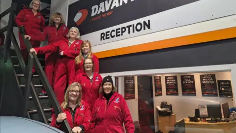 Davanti Tyres Seven women wearing red overalls stand on a staircase. There is a white wall behind them and a window partition, which shows a reception area through it