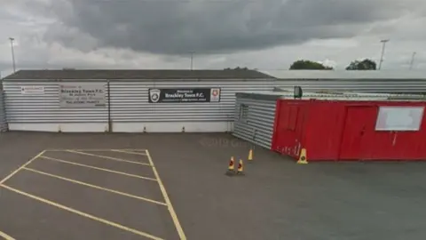 Google Single-storey corrugated iron buildings, mostly grey but with one red painted wall to the right.  A sign says "Brackley Town FC".