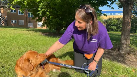 Rhubarb, a golden-brown assistance dog wearing a harness and “school assistance dog” lead, stands on grass as Zoe Latter crouches beside him holding the lead, with trees and college buildings in the background. She is wearing a purple T-shirt with a logo that says "Team Rhubarb".