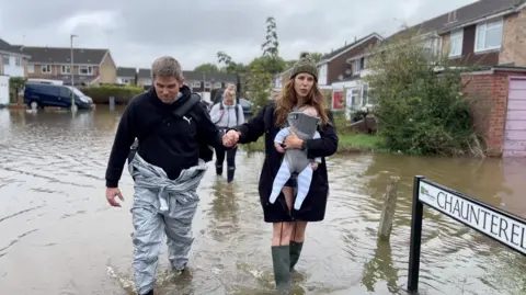 A family of three walks through flooded water in their street, Chaunterell Way, there are homes in the background and a black van to the left of the frame, the dad is standing to the left, he has short hair and wears a black hoodie with a Puma logo on the right of his chest and a silver waterproof boiler suit. He is holding a woman's hand, she wears a khaki green woollen beanie hat with a khaki green fur bobble on top of it and a black parka coat and green wellie boots, she carries a baby in a baby carrier