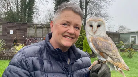 Sharon Cox, who has short white hair and is wearing a black puffer jacket, smiles at the camera. A small owl is perched on her gloved left hand and also looking towards camera. Over Sharon's shoulder a large black bird of prey is stood on a metal fixture in front of an aviary. 