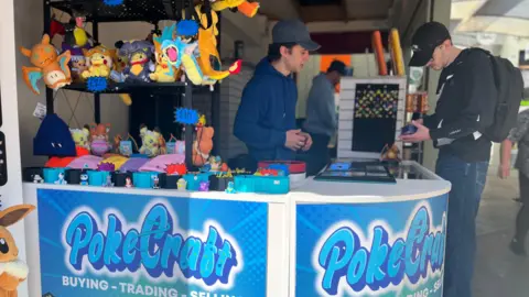 Jack Maclean/BBC A market stall has blue branding on it and lots of toys on the counter and shelves behind. A man in a blue jacket is serving a customer who is wearing a black top.