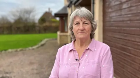 A grey-haired woman with a pink blouse standing outside a large country house