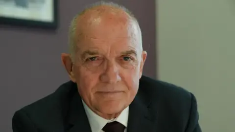 Colin Anderson looking directly at the camera. He has grey hair and is wearing a black suit, white shirt and black tie.
