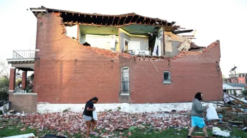 Reuters Bricks from a collapsed wall can be seen on the ground as two residents carry belongings away from a heavily damaged home in St Louis, Missouri, on 16 May 2025. The roof and several rooms on the upper floor are damaged and exposed.