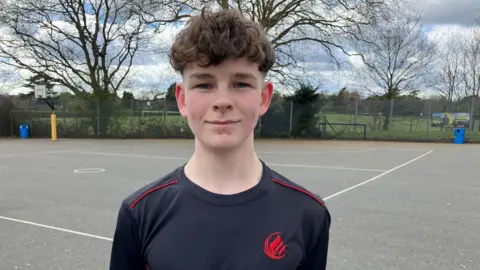 Oscar is stood in the school playground in a school branded black top with red piping and a red logo. He has mousy coloured, curly hair, and his hands behind his back.