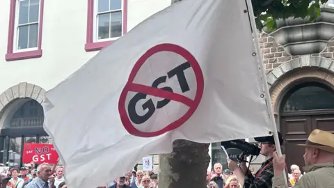 A crowd of people gather in a town square. In the foreground is a white flag containing a red circle with a red line through it, like a traffic sign. The letters "GST" are scored through by the line