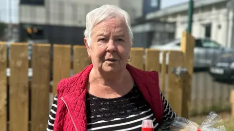 Elsie Ferris has short grey hair and is wearing gold hoop earrings. She has a red gilet jacket on and is wearing a black white stripped long sleeve top. In her hands is the groceries she bought from the shop.