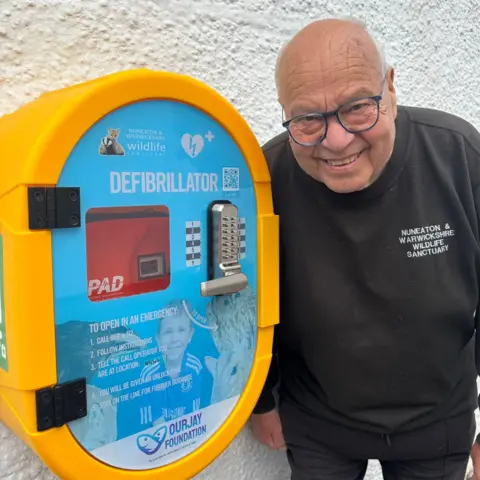 Nuneaton & Warwickshire Wildlife Sanctuary Geoff Grewcock stands next to a defibrillator fitted on the wall of his wildlife sanctuary and smiles. He wears a black jumper with the sanctuary's name on. The defibrillator case has a picture of a young Jamie on it, surrounded by animals.