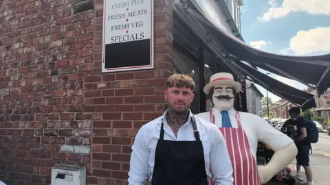 A younger man with tattoo on this neck, wearing a white shirt and a black apron, stands outside a red-brick butchers shop. A model butcher, with a hat, striped red apron and extravagant moustache, stands behind him.