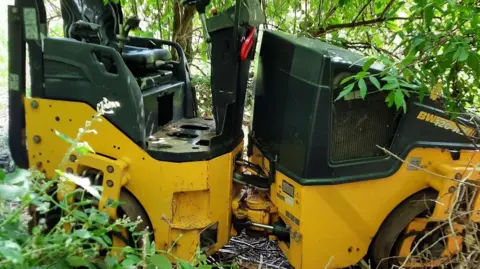 A large yellow and green road roller is in a hedge.