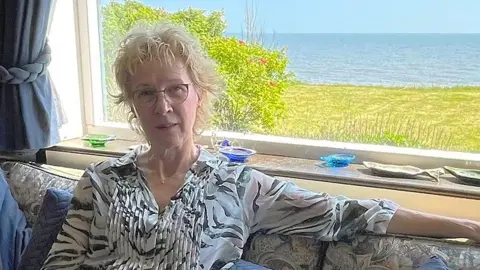 A woman is sitting on a sofa in front of a window. A strip of garden can be seen outside and beyond it the sea.