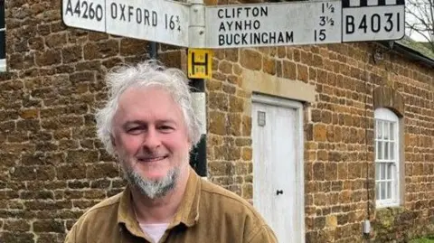 Gareth Epps, a man with white hair and a beard, stands in front of a stone house and smiles for the photograph. He is wearing a brown collared shirt over a white T-shirt. Directly behind him is a sign that gives various locations - including Oxford and Buckingham - and their distance away in miles. 