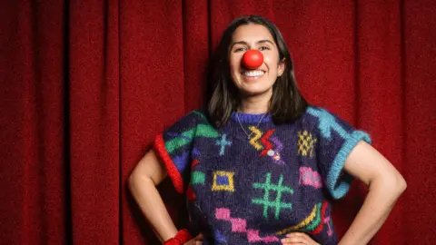 A woman with shoulder-length dark hair and wearing a short-sleeved jumper patterned with multicoloured symbols, and a fake red nose. She's smiling at the camera with a red curtain behind her