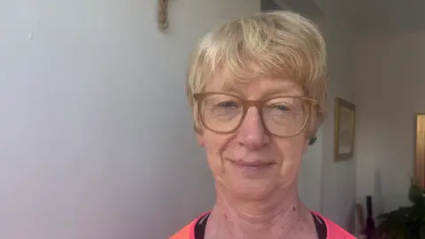 Maureen is standing on her doorstep looking at the camera and smiling. She has short brown hair and glasses and a pink top on.