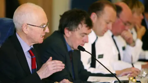 NI Policing Board Sir Desmond Rea sitting at a table in front of a microphone with a row of other people sitting beside him. He is bald, wearing glasses, a black blazer, pink shirt and red tie.