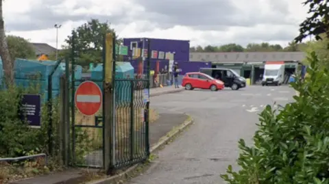 An entrance to a recycling centre with a metal gate, surrounded by hedges and a red car and two vans parked in front of large purple blocks behind that