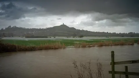 Mike Jefferies Dark storm clouds hover over Glastonbury Tor in the background. In the foreground the river has burst its banks, with water covering a number of fields. 