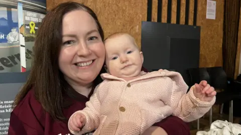 Women with dark hair in red jumper holding baby who is wearing a pink outfit. 