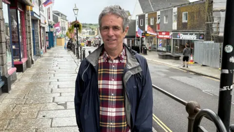 BBC A man with grey hair wearing a black, white, and yellow plaid shirt and dark blue rain jacket with Market Jew Street in the background. 