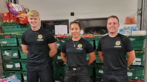 Two men and a woman in black tshirts and trousers standing in front of a large stack of green crates containing a variety of items