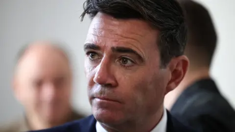 Andy Burnham wearing a black suit and white shirt, looking to the left of the frame of the photograph. The picture is a close up with most of Burnham out of shot bar his head and shoulders.