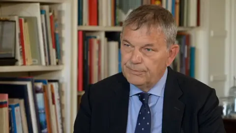 Philippe Lazzarini sits in front of a white bookshelf. He has greying hair and wears a blue shirt and dark jacket. He also wears a navy tie with small grey embellishments.