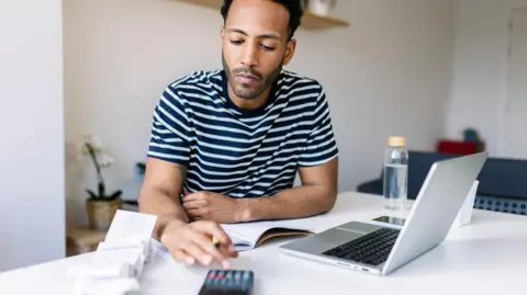 Getty Images Man sits at a desk with a laptop computer and a calculator as he looks at paperwork