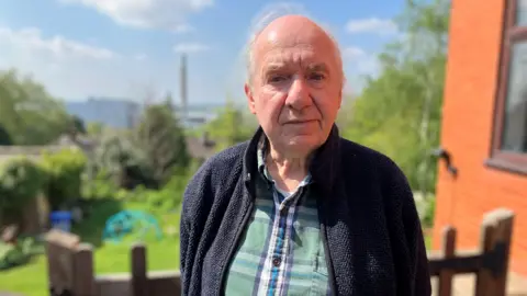 Ralph Owen is standing in the back garden of his home and he is staring directly at the camera. Behind the grass and bushes in his garden, you can see the incinerator.