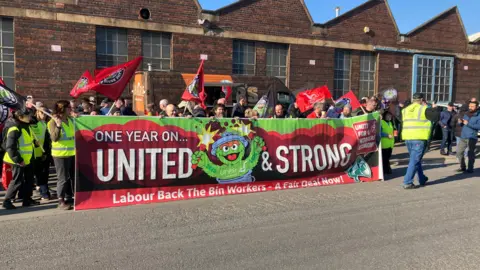 A large group of people in high-visibility vests and dark clothing stand behind a wide red and green banner reading 'One Year On... United & Strong'. Several red Unite union flags are being waved above the crowd. The group is gathered on tarmac in front of a brick industrial building with large windows on a bright, clear day.