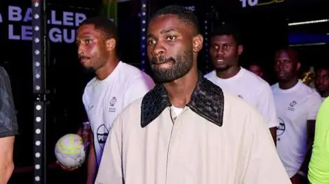 Getty Images Dave walks out onto the pitch at London's Copperbox Arena wearing a cream jacket with black collar. Three players walk in behind him wearing white t-shirts