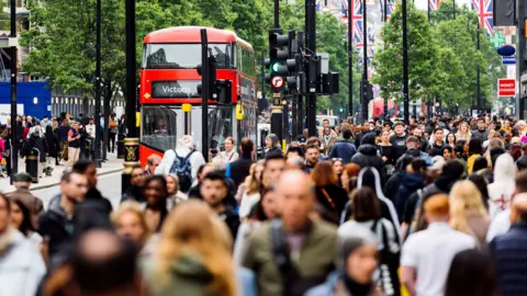 Getty Images A very busy shopping street filled with people with a red London bus on the road. There are trees, Union flags and traffic lights visible in the background. 