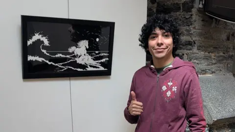 JUAN ASHLISH Juan Ashlish, who has dark curly hair and is wearing a dark pink hoodie and it making a thumbs up gesture. he is standing next to a black and white painting of Manannan's horse Enbarr