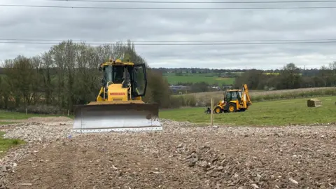 BBC A yellow bulldozer faces the camera with its silver scraper at the front. It sits on a large area of soil that is amid a grassy field, and the soil appears to have been recently turned over. There are trees and fields in the background.