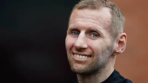 Rob Burrow has short brown hair and is smiling looking into the camera. He is wearing a black top.