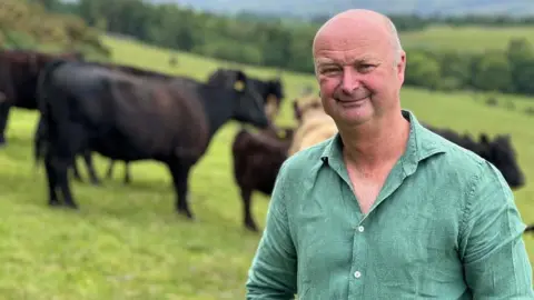 John Renner smiling into the camera. He is standing on a field in front of some black cows which are out of focus. He is wearing a green shirt and has balding, grey hair.