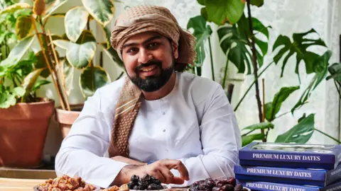 Date Sultan Shah wears a traditional white dress and patterned brown turban and poses at a table with dates on it. Behind him are plants