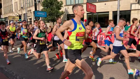 Athletes running through Bristol City Centre for the AJ Bell Great Bristol Run. They are all wearing running vests, race bibs, shorts and running shoes.